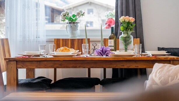Ett dukat matbord med tallrikar, glas och en flaska, med utsikt genom fönstret mot en byggnad och en vas med blommor.