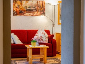 Ett vardagsrum med en röd soffa, ett litet träbord med en växt och en tavla på väggen.