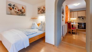 Ein Schlafzimmer mit Bett, Kissen und Wandbildern. Eine Küche ist durch einen Bogen sichtbar.