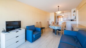 A modern living room with a flat-screen TV, blue sofas, a dining table with chairs, and a kitchen area in the background. - Apartment on the 6th floor with sea view (Middelkerke)