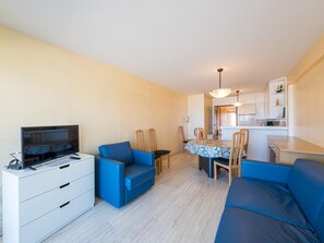 Ein modernes Wohnzimmer mit Flachbildfernseher, blauen Sofas, einem Esstisch mit Stühlen und einer sichtbaren Küchenzeile im Hintergrund.