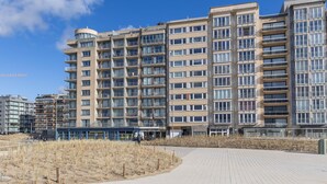 A modern multi-story residential building with balconies, a covered walkway, and a sandy area in front. - Apartment on the 6th floor with sea view (Middelkerke)