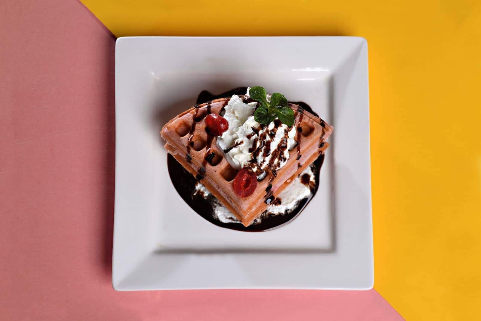 A square white plate with a dessert on a yellow and pink background.