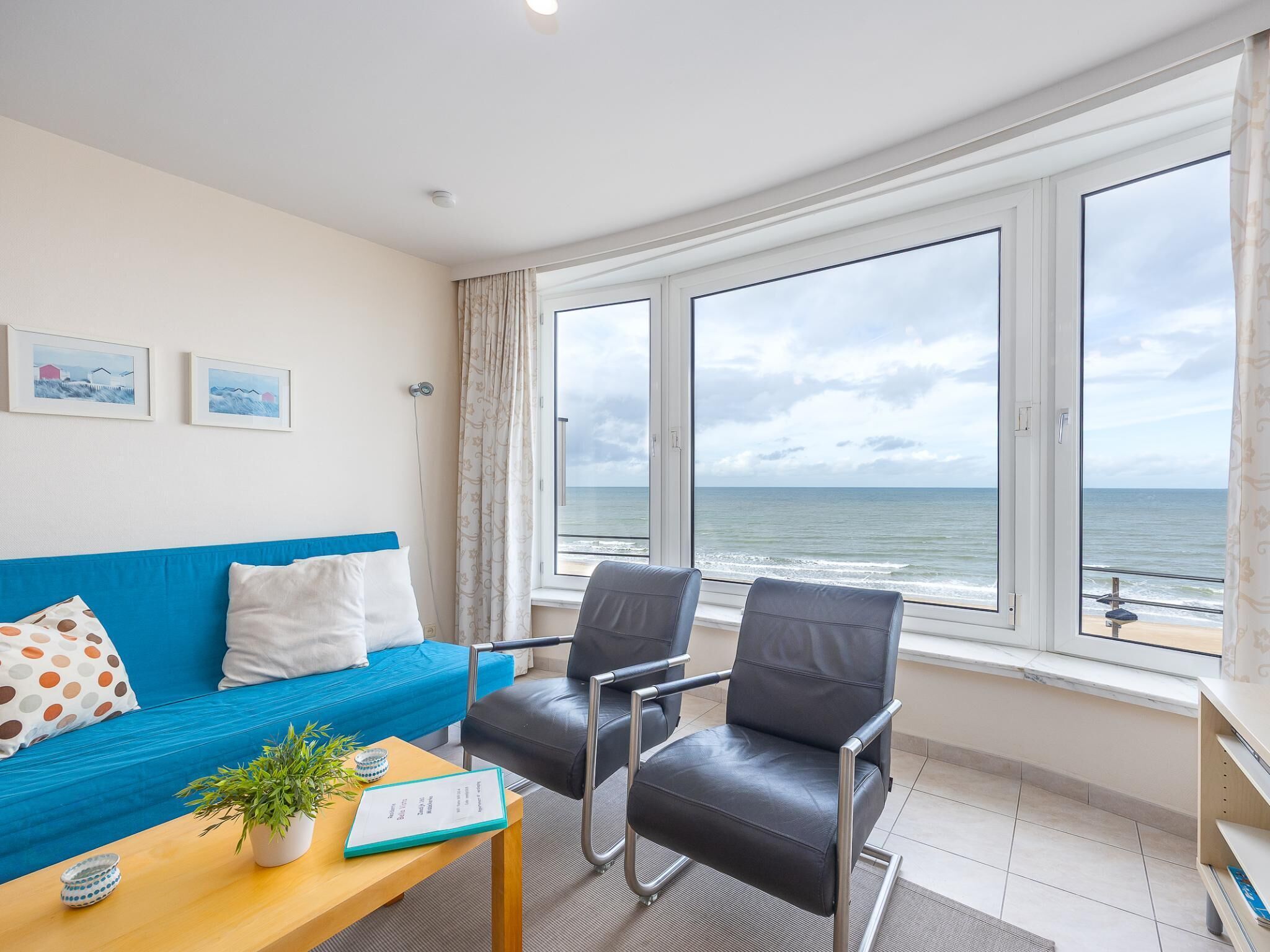 Une pièce moderne avec un canapé bleu, deux fauteuils noirs, une table en bois ornée d’une plante et d’un carnet, et de grandes fenêtres donnant sur la mer.