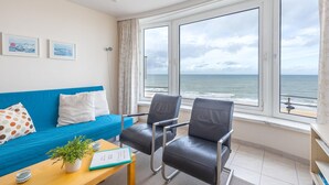 Ett modernt rum med en blå soffa, två svarta stolar, ett träbord med en växt och ett anteckningsblock samt stora fönster med utsikt över havet.