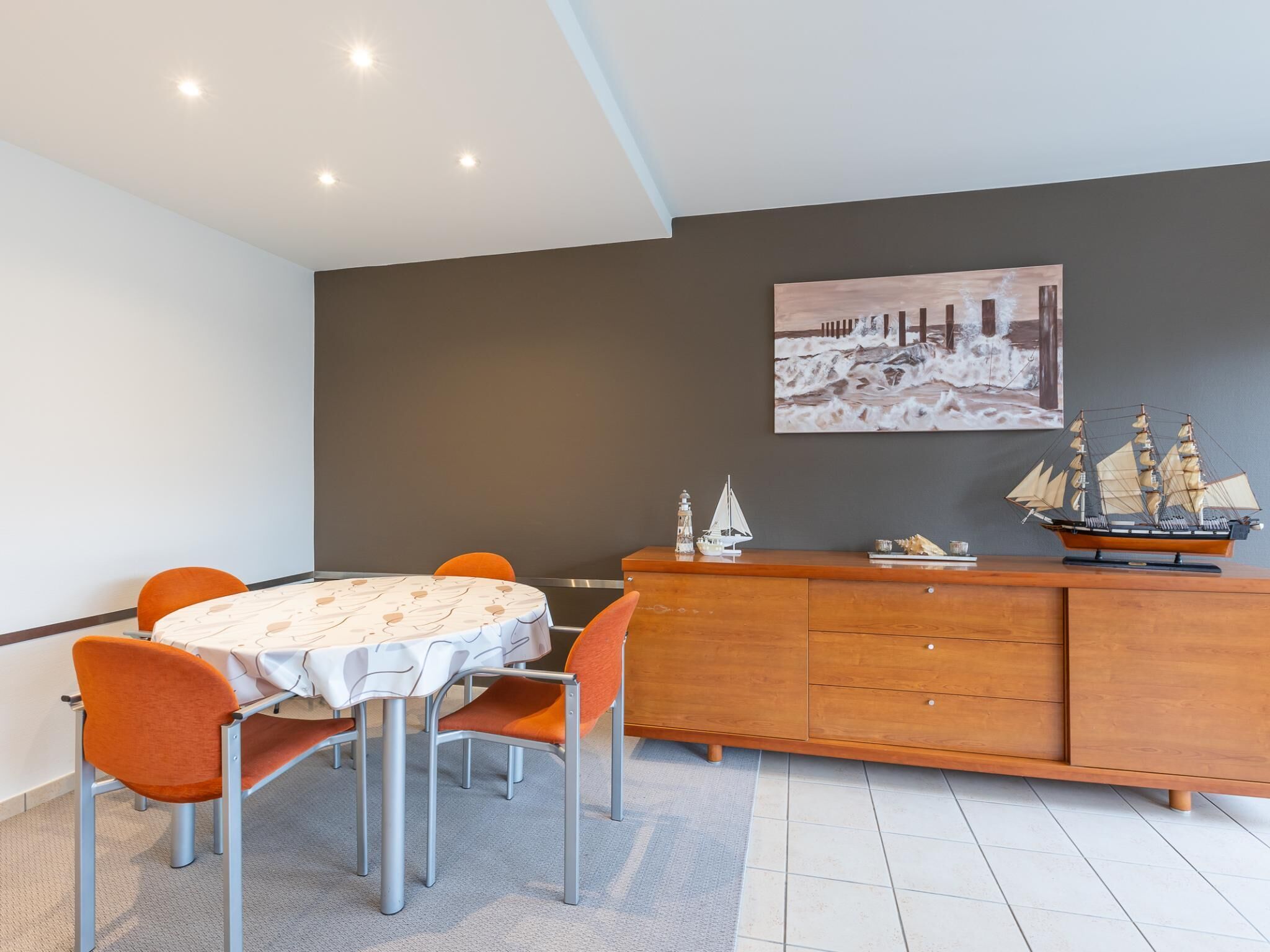 A dining area with a round table, orange chairs, a wooden cabinet, and a wall-mounted painting.