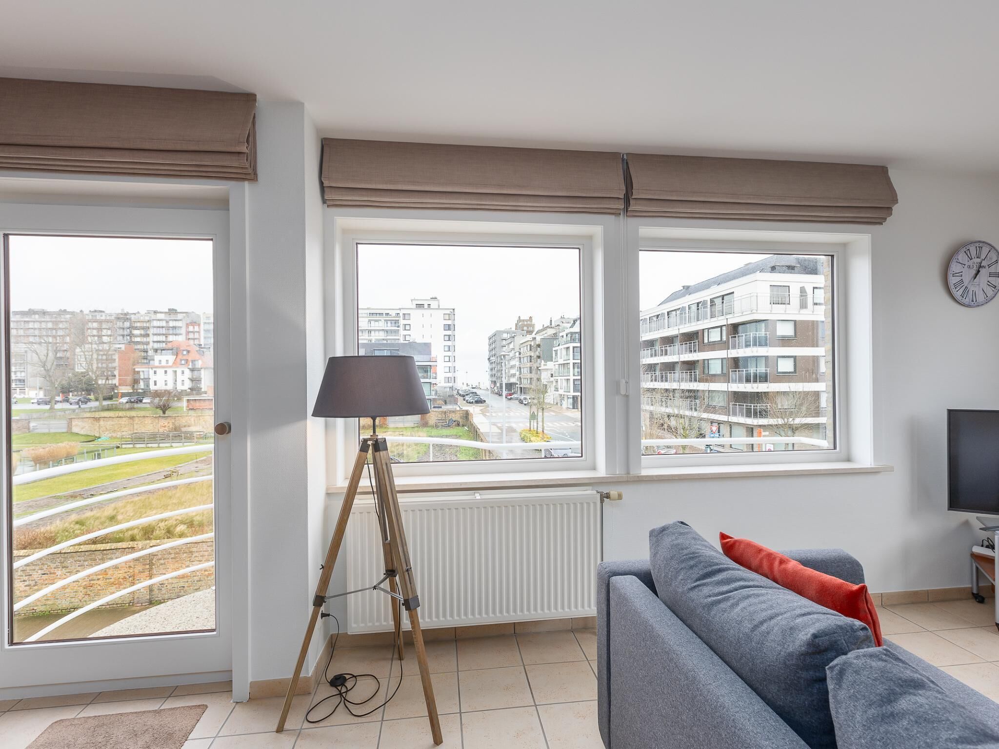 Een moderne woonkamer met een bank, een lamp en een televisie. Drie ramen bieden uitzicht op gebouwen en een park.