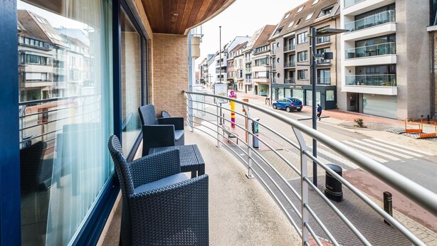 A balcony with wicker chairs, a view of modern buildings, and a street with a parked car. - Apartment with 2 bedrooms and large terrace (Middelkerke)