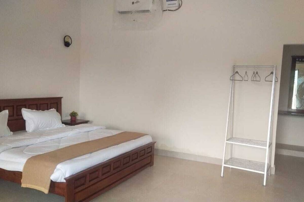 A bedroom with a wooden bed, a white bedside table, a small potted plant, a white metal shelf, and a mirror above the shelf.