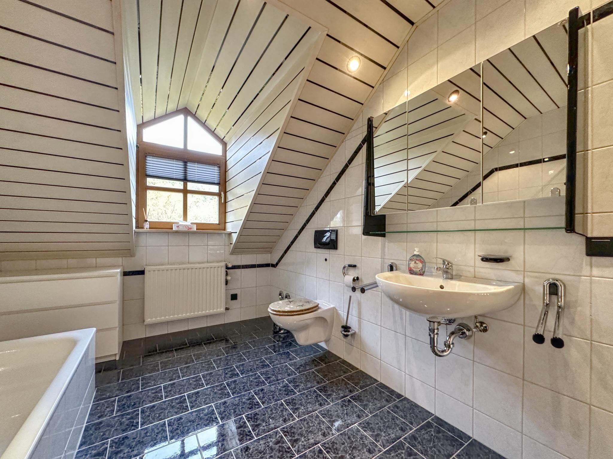 A modern bathroom with a sloped ceiling, a large window, a bathtub, a toilet, and a sink.