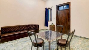A room with a brown leather sofa, a glass-top table with two chairs, a wooden door, and a window with blue curtains.