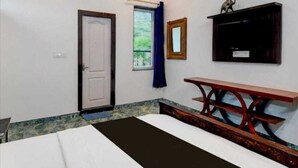 A room with a bed, a window with a curtain, a door, a small shelf with a mirror, and a wooden bench.