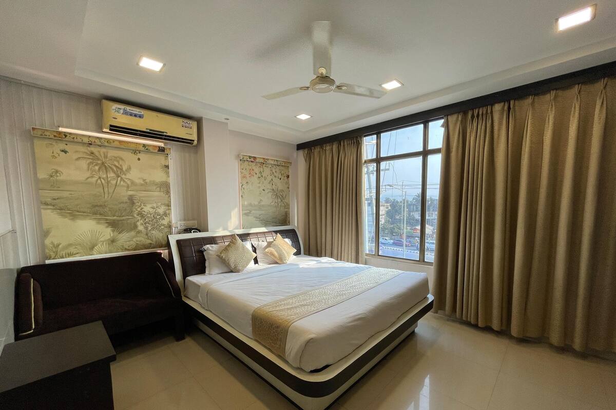 A modern hotel room with a large bed, a ceiling fan, an air conditioning unit, and a view of the city through large windows.