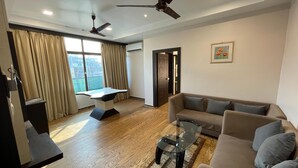 Ein modernes Wohnzimmer mit einer Couch, einem Glastisch und einem Schreibtisch mit Stuhl. Zwei Deckenventilatoren und ein großes Fenster mit Vorhängen sind vorhanden.