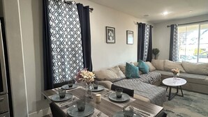 A modern living room with a dining area. There is a sofa with cushions, a coffee table, and a dining table set with plates, glasses, and cutlery. The room has a patterned curtain and a ceiling light. - Spacious 4-bedroom house in charming Newark with AC (Newark)
