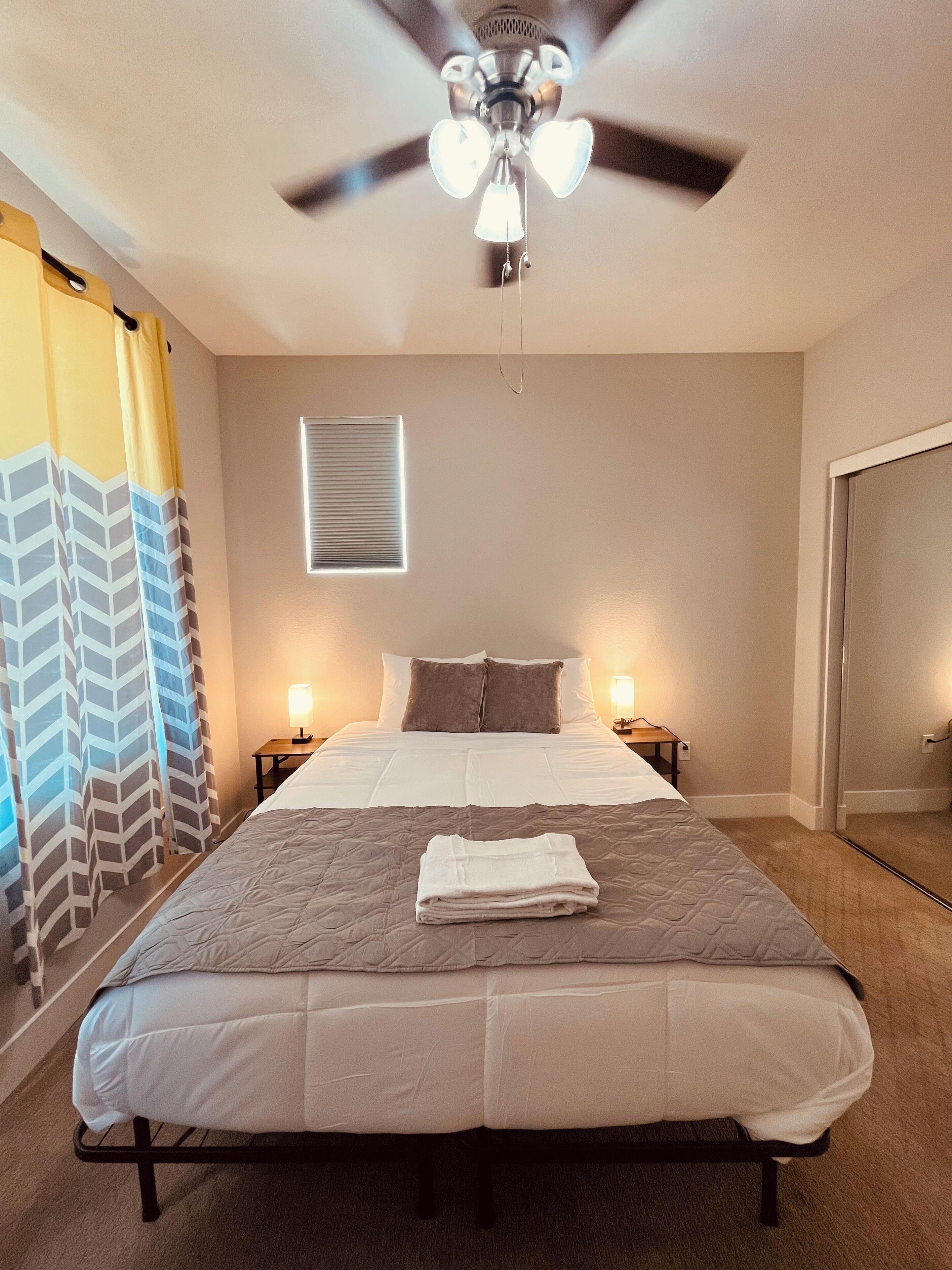 A neatly made bed with a folded towel, a ceiling fan, a window with blinds, and a nightstand with a lamp.