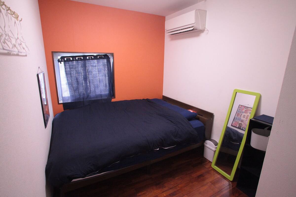 A small bedroom with a single bed, an air conditioner, a window with curtains, and a mirror.