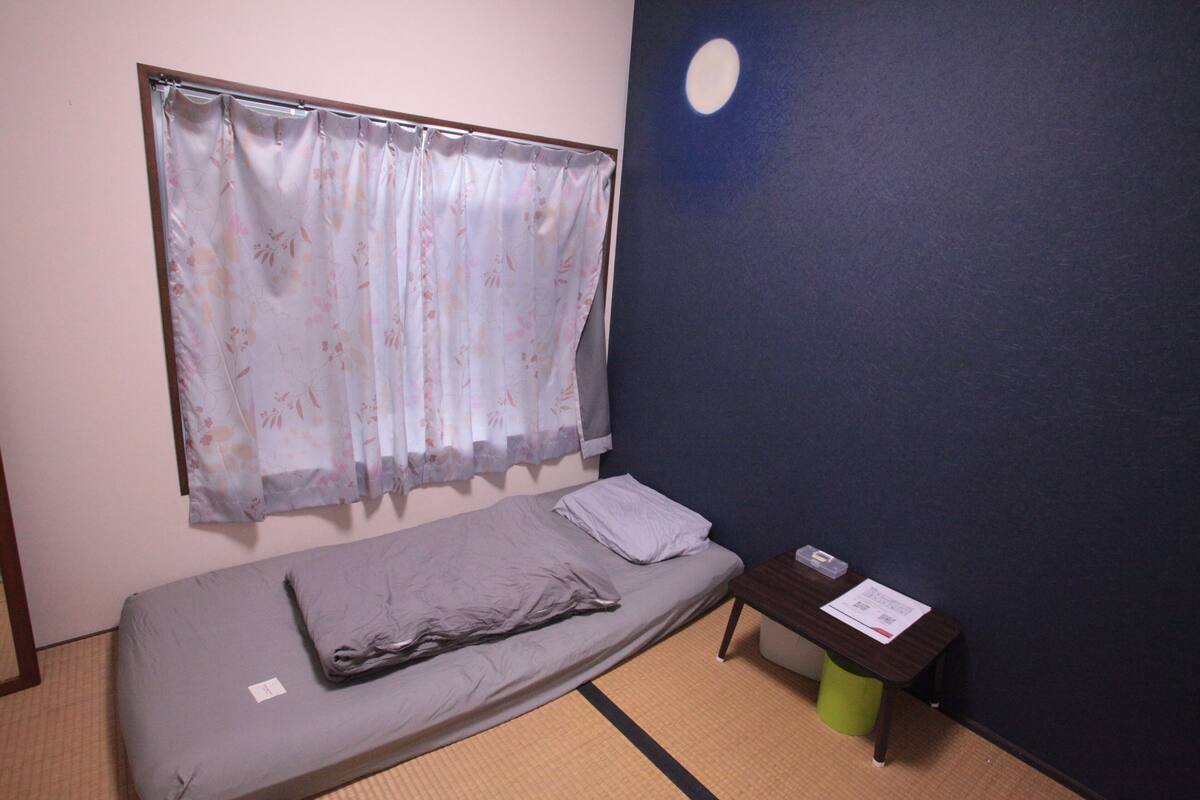 A single bed with a floral curtain, a small table with a green trash bin, and a wall with a moon painting.