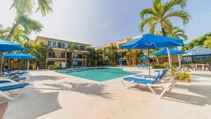 A pool area with blue umbrellas and lounge chairs, surrounded by palm trees and a building with balconies. - Silent residence in downtown Sosua, you don't hear that every day! (Sosúa)