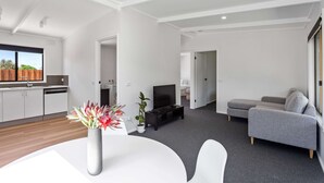 Ein modernes Wohnzimmer mit grauem Sofa, einem Flachbildfernseher und einem Essbereich mit weißem Tisch und Blumenvase.