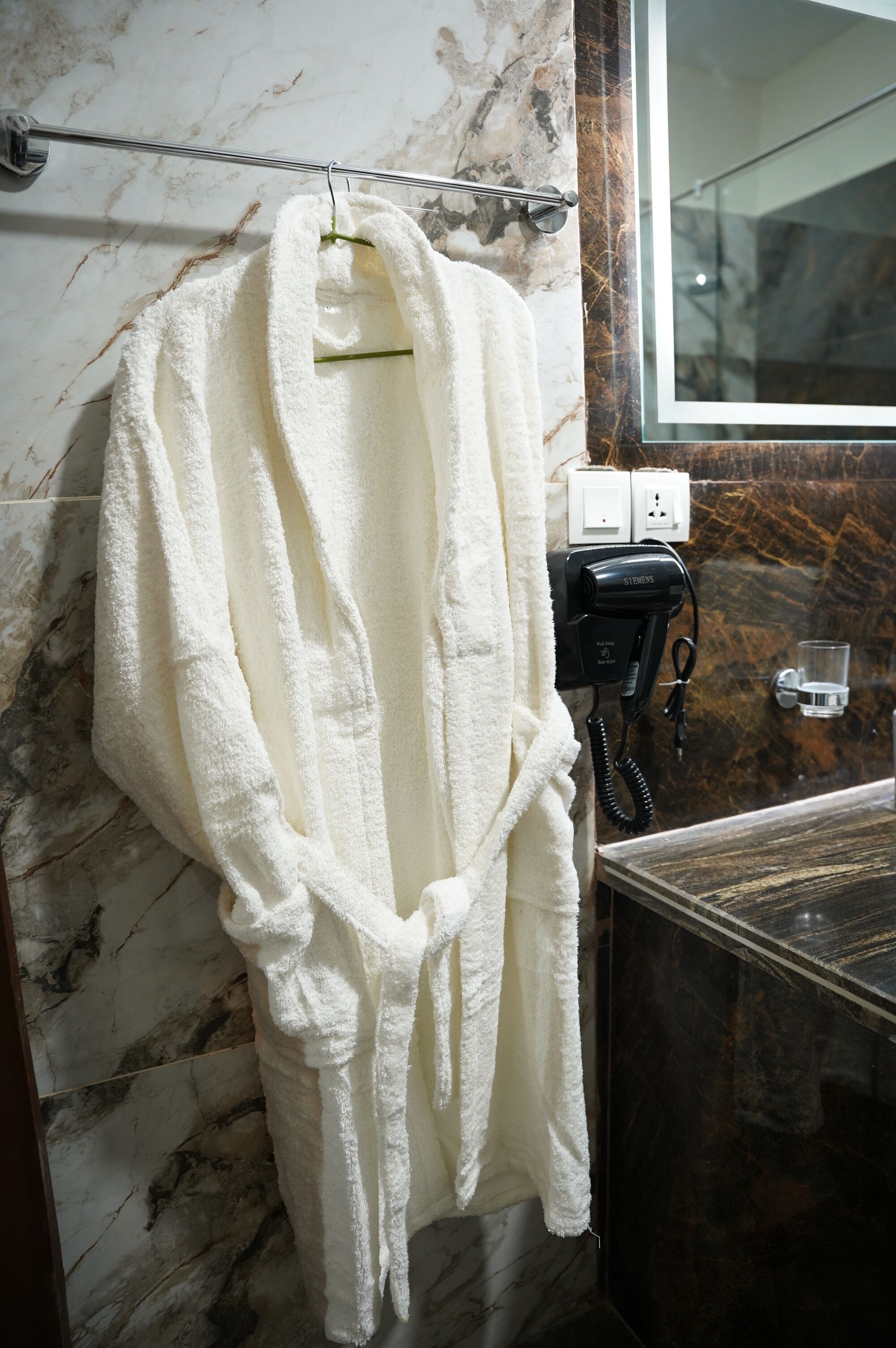A white robe hanging on a rack in a bathroom with marble walls and a glass shower enclosure.