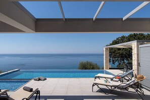 Ein Poolbereich mit freiem Blick auf das Meer, Liegestühle und eine überdachte Sitzgelegenheit.