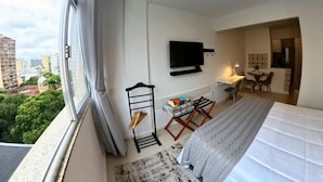 Une chambre d’hôtel moderne dotée d’une grande fenêtre, d’un lit, d’un bureau avec une chaise, d’un téléviseur fixé au mur et d’un petit coin cuisine.