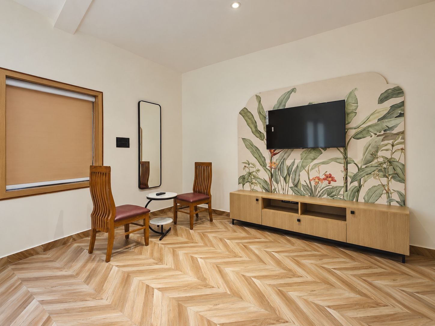 Una habitación con suelo de espiga, un televisor montado en una pared con papel tapiz tropical, dos sillas de madera, una mesita redonda y una ventana con persiana.