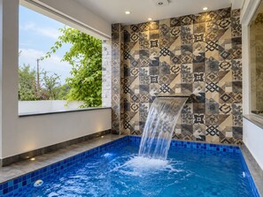A modern indoor pool with a waterfall feature, surrounded by patterned tiles and a window with a view of greenery. - The Forest Hills Resort (Sarada)