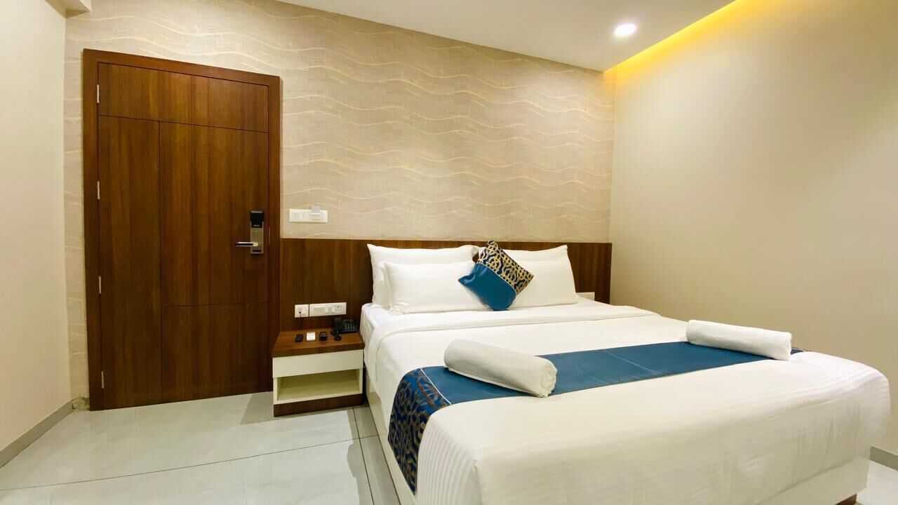 A hotel room with a large bed, a wooden wardrobe, a nightstand, and a wall-mounted light fixture.