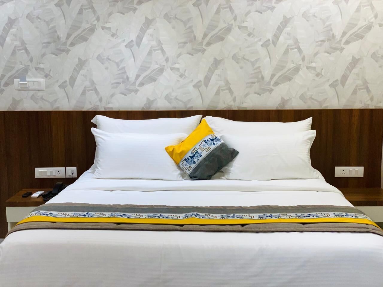 Una cama bien hecha con ropa blanca y una colcha estampada, una mesita de noche con un teléfono y un control remoto, y una pared con papel tapiz de diseño floral.