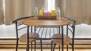 A round wooden table with a woven basket containing snacks and bottled water, surrounded by metal chairs, against a window with curtains. - Fish, Hike & Explore: Mtn-View Lake George Studio (Lake George)