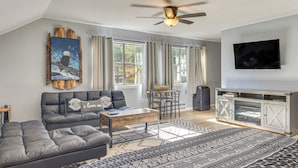 A modern living room with a sectional sofa, a coffee table, a TV, and a large window with curtains. - Fish, Hike & Explore: Mtn-View Lake George Studio (Lake George)