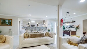Ein geräumiges Wohnzimmer mit einem weißen Sofa, einem Couchtisch mit Schachbrett und einer Küche im Hintergrund.