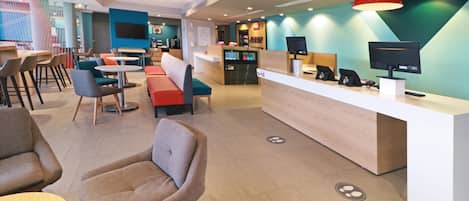A modern hotel lobby with a reception desk, seating areas, and a large TV screen.