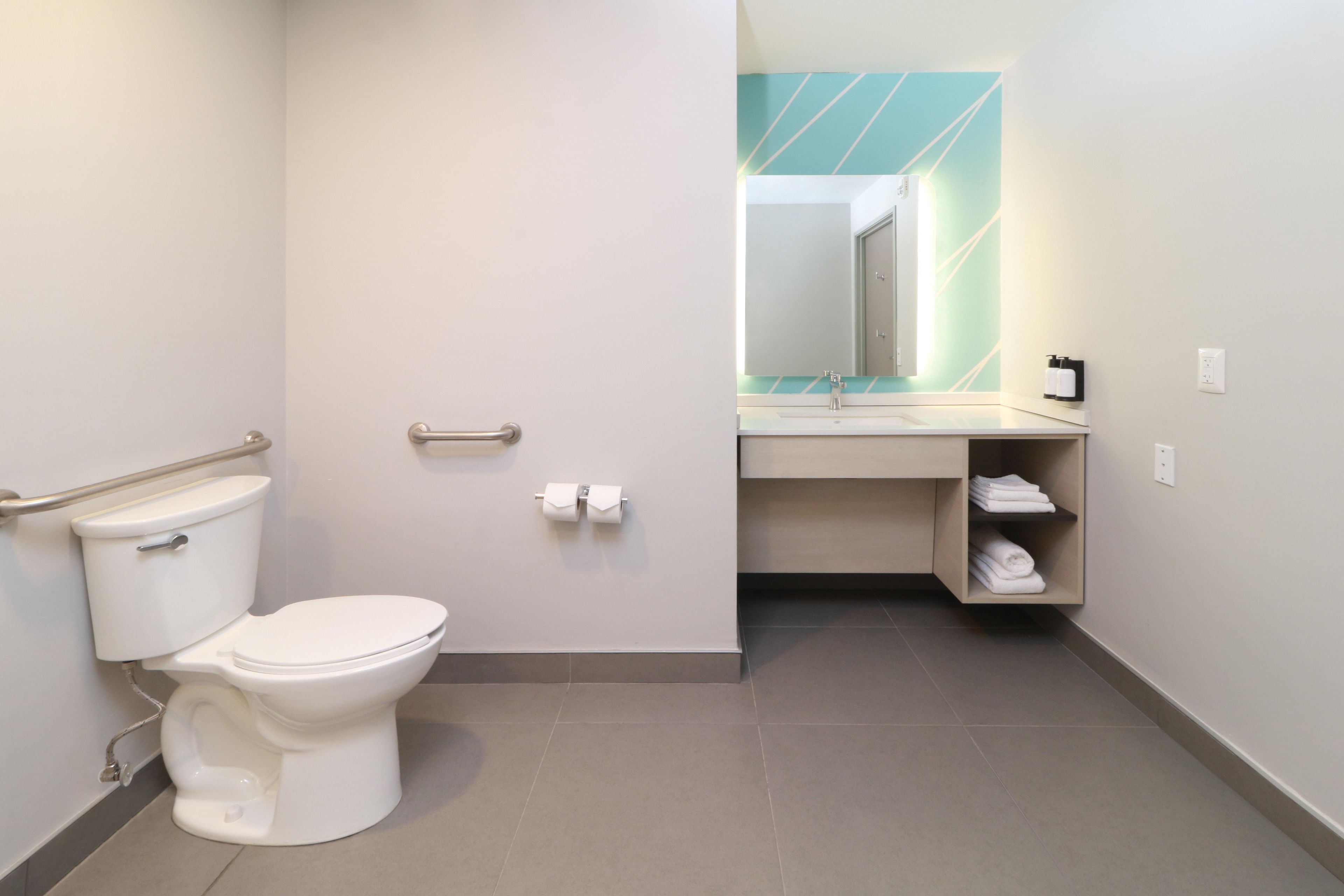 A modern bathroom with a toilet, handrail, sink, mirror, and towel storage.
