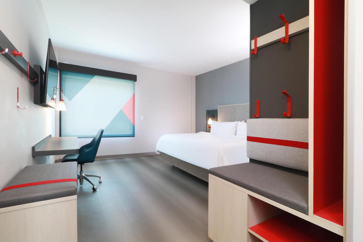 A modern hotel room with a bed, desk, chair, and built-in TV.