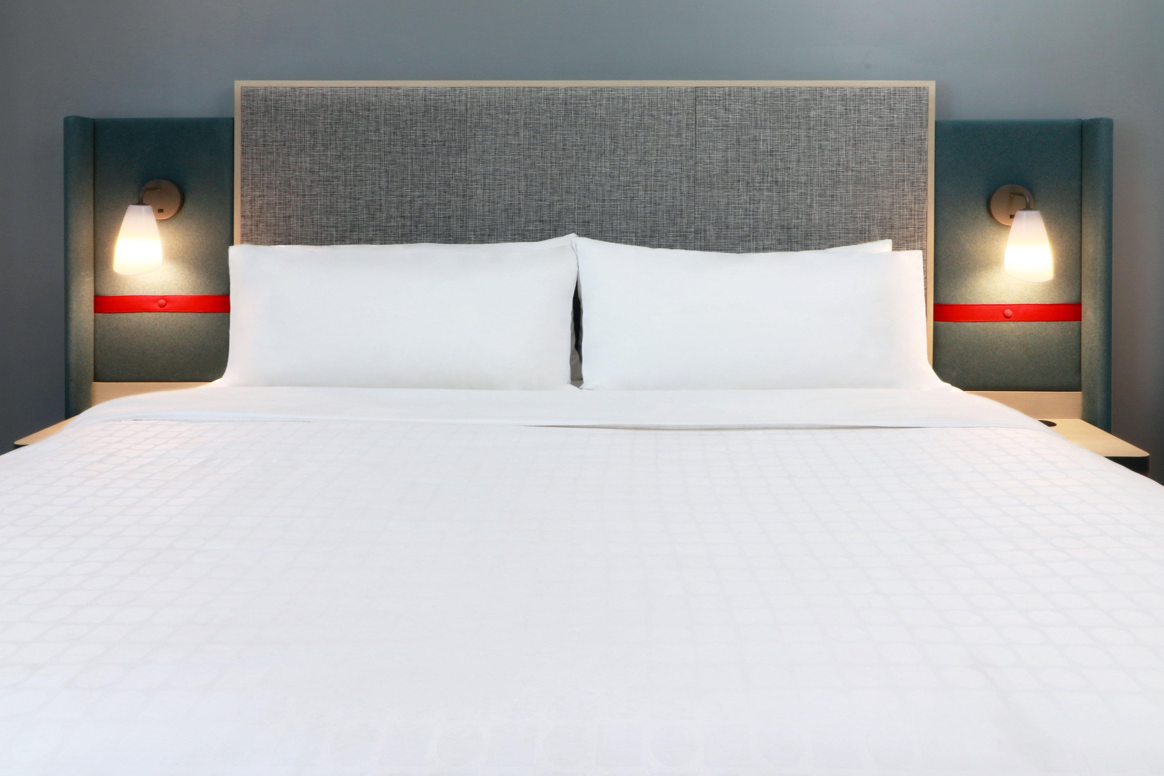 Una cama con cabecera gris, dos mesitas de noche con lámparas y un detalle rojo en las mesitas.