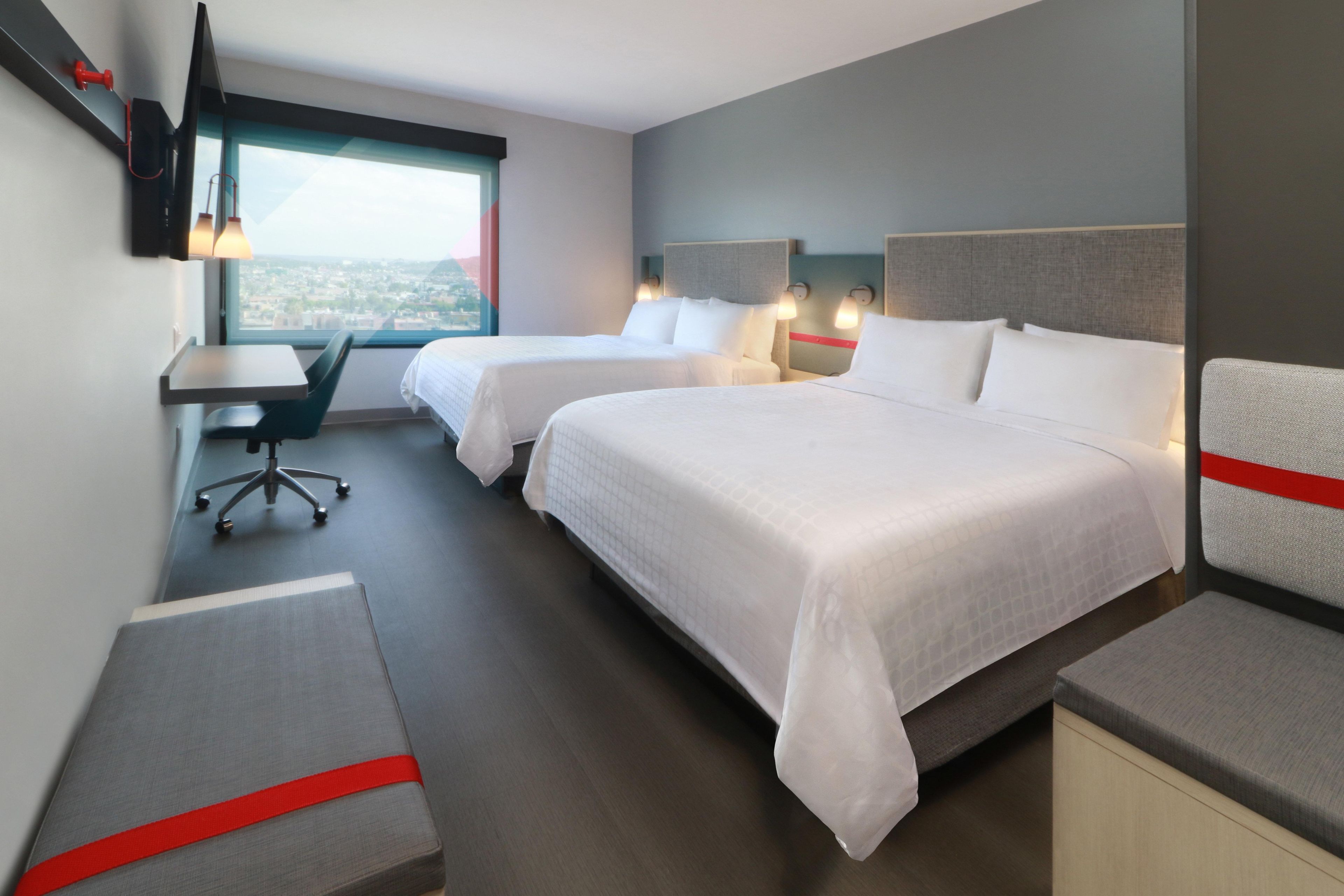 A hotel room with two beds, a desk, a chair, and a TV.