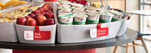 Esposizione di prodotti alimentari tra cui mele e yogurt in contenitori etichettati 'Fruit burst' e 'Classic grains'.