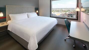 Habitación de hotel con cama, mesitas de noche, escritorio y un amplio ventanal con vistas a la ciudad.