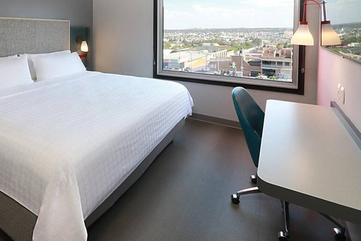 A hotel room with a bed, a desk, a chair, a nightstand, and a window with a city view.