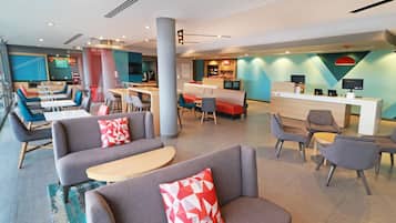 A modern hotel lobby with seating areas, a reception desk, and a dining area.