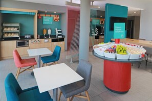 A modern café with a central counter displaying food items, seating with colorful chairs, and a kitchen area with appliances. - Avid Hotel Fresnillo (Fresnillo)