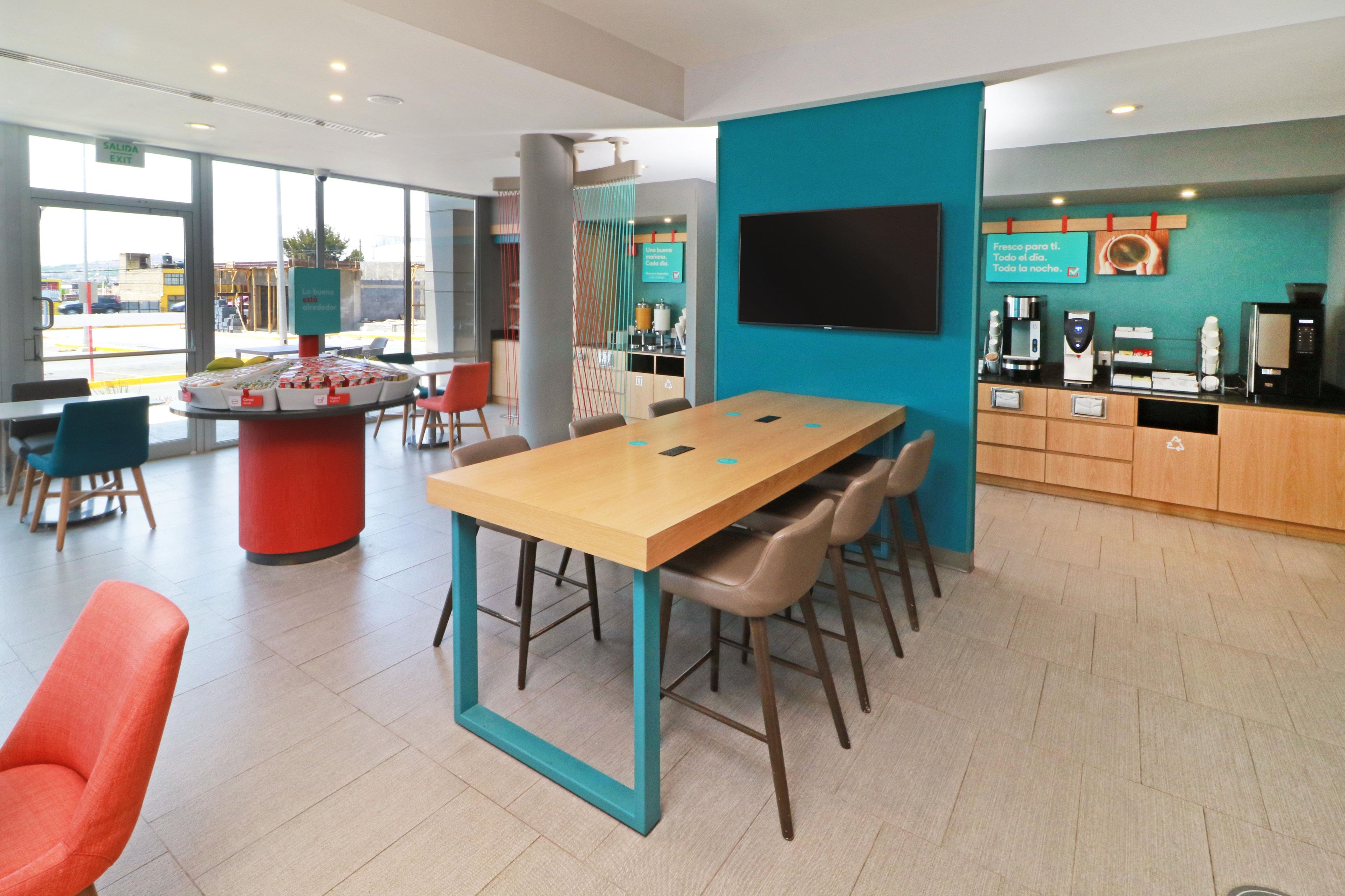 A modern hotel lobby with a large wooden table, comfortable seating, a coffee station, and a TV mounted on the wall.