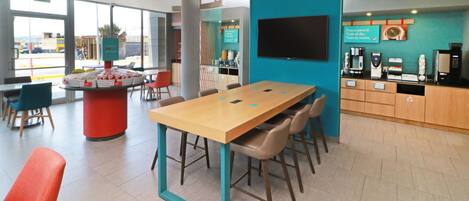 A modern hotel lobby with a large wooden table, comfortable seating, a coffee station, and a TV mounted on the wall.