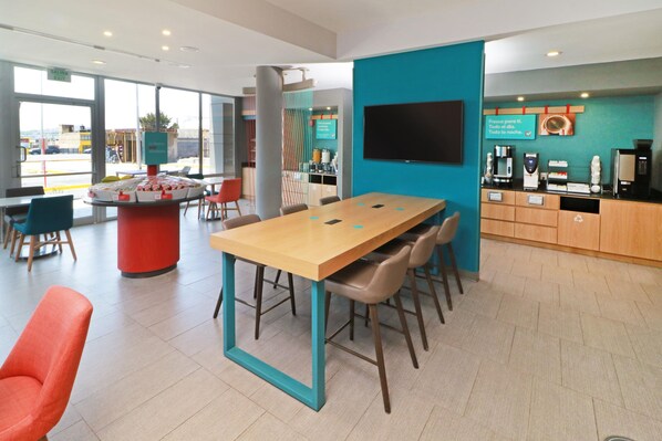 A modern hotel lobby with a large wooden table, comfortable seating, a coffee station, and a TV mounted on the wall.