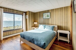 Een slaapkamer met een groot raam, houten wanden, een bed, een nachtkastje en een lamp.