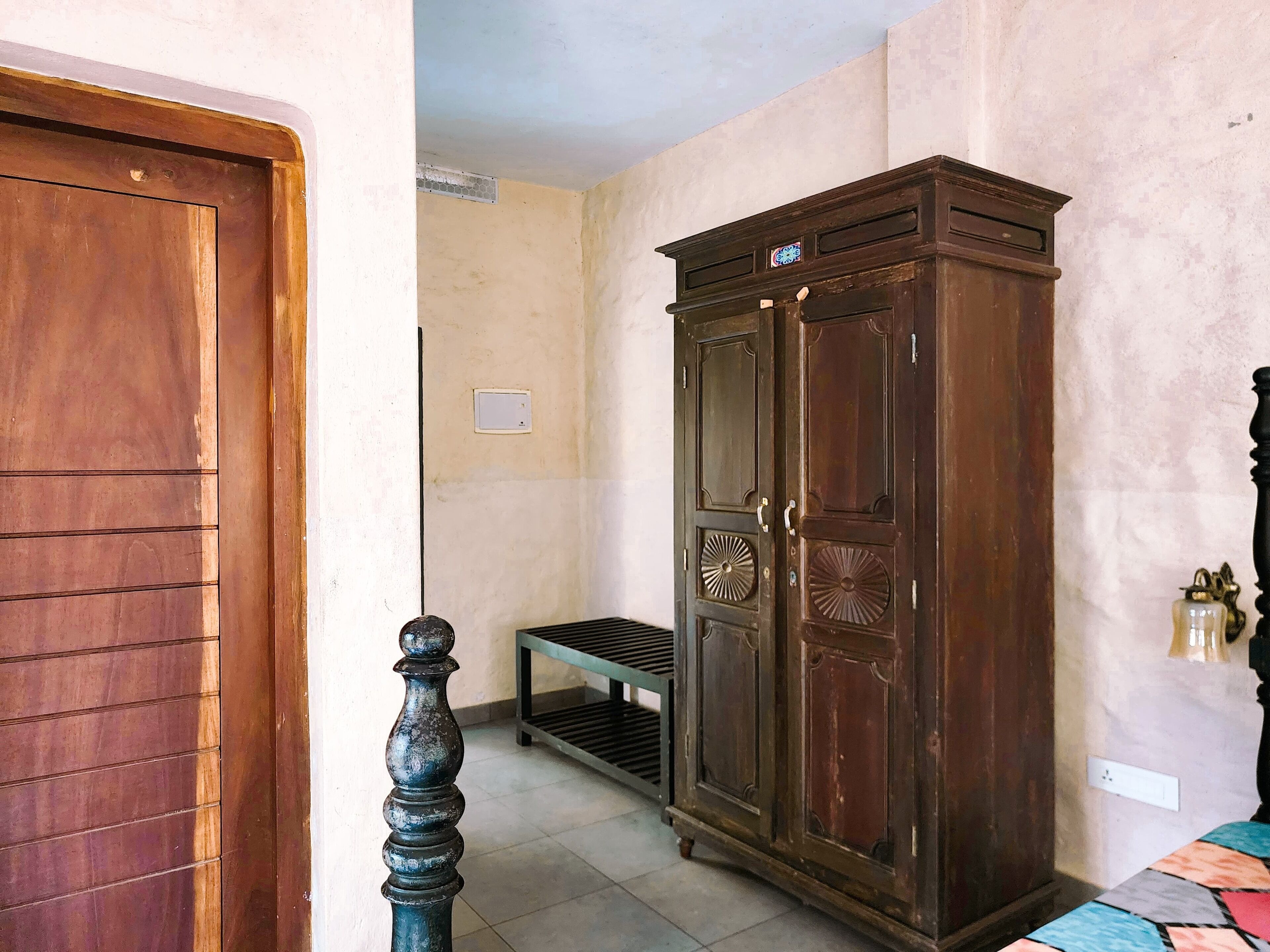 Una habitación con puerta de madera, un armario de madera oscura, una mesita y una cama con colcha estampada.