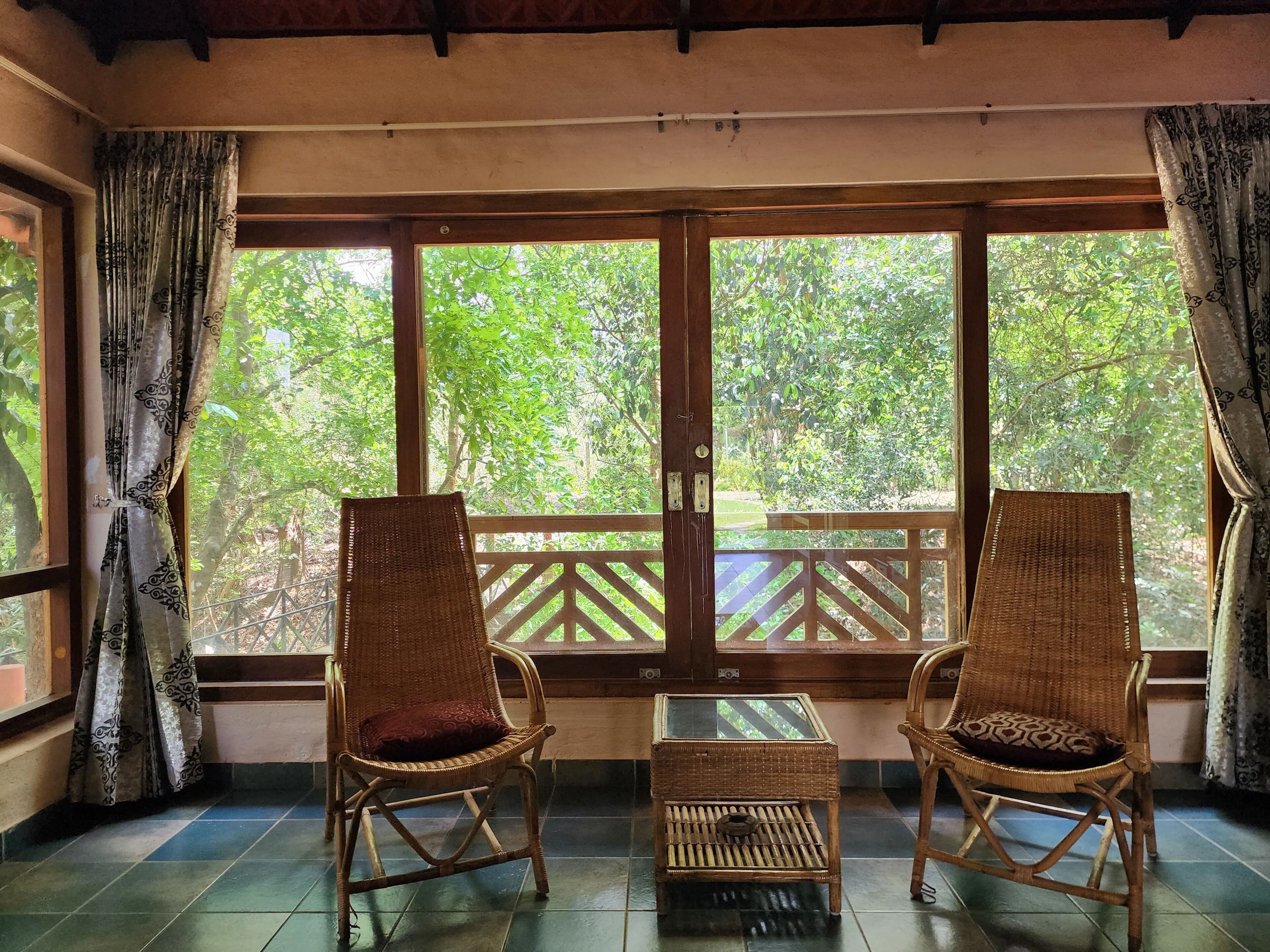 A room with two wicker chairs and a small table, large windows with a view of greenery, and a wooden balcony railing.